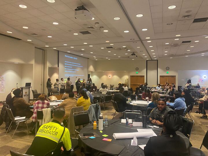 A wide view of a conference room where participants sit at round tables while a group presents at the front beside a large projection screen.