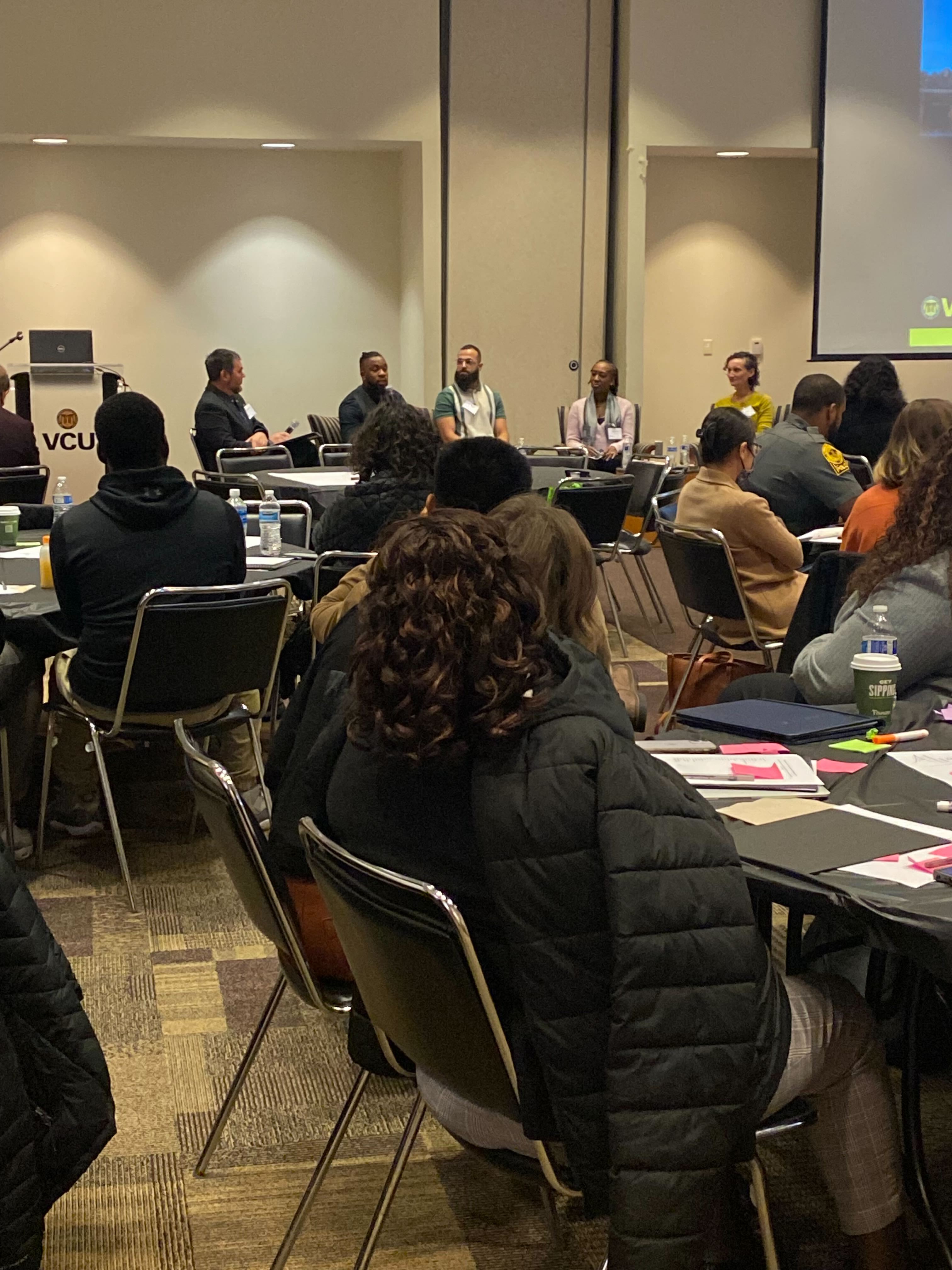 A closer view of conference attendees seated at tables watching a panel of speakers at the front of the room near a large presentation screen.