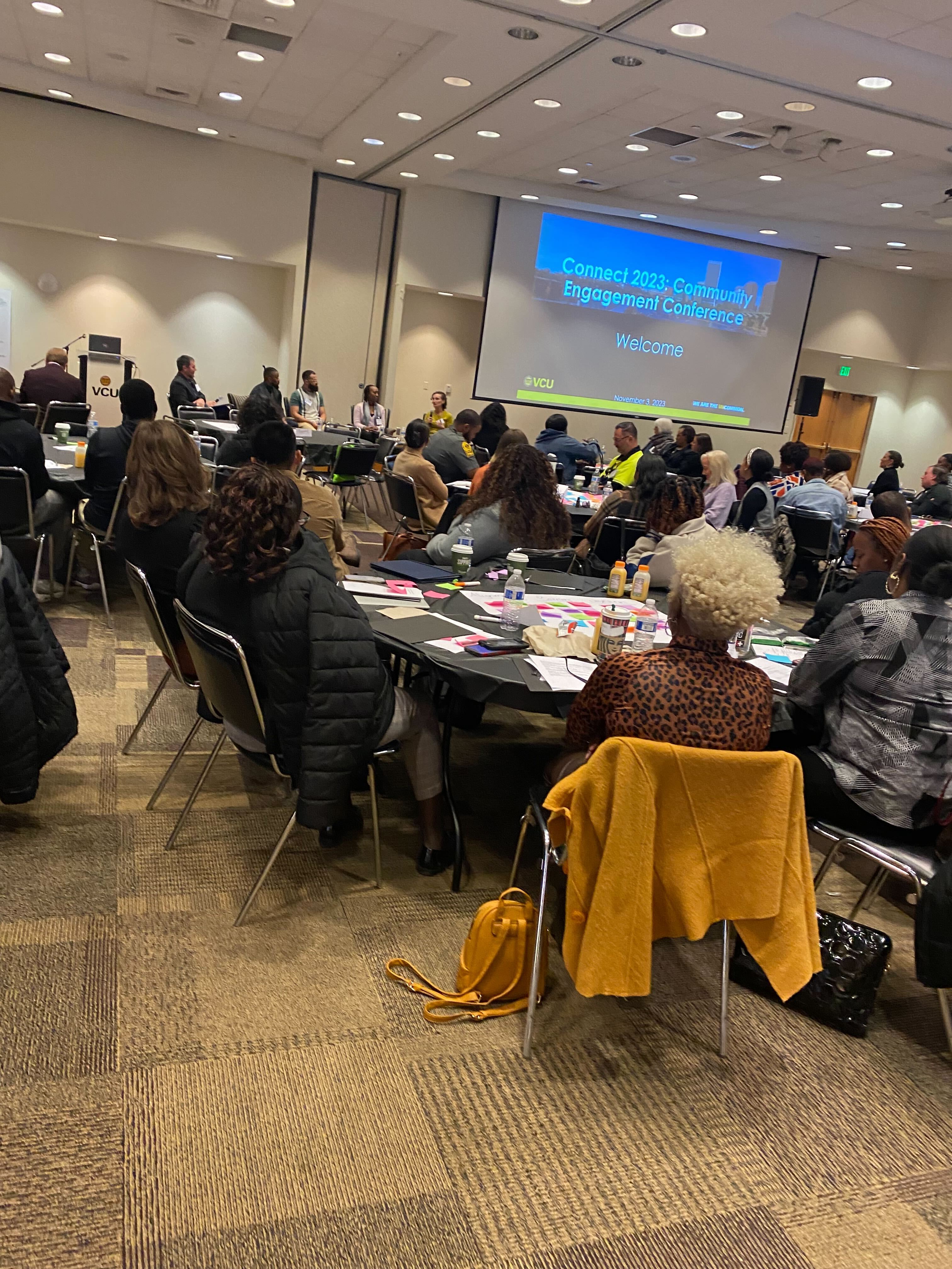 Conference attendees sit at round tables facing a large screen displaying the title “Connect 2023: Community Engagement Conference” as a panel speaks at the front of the room.