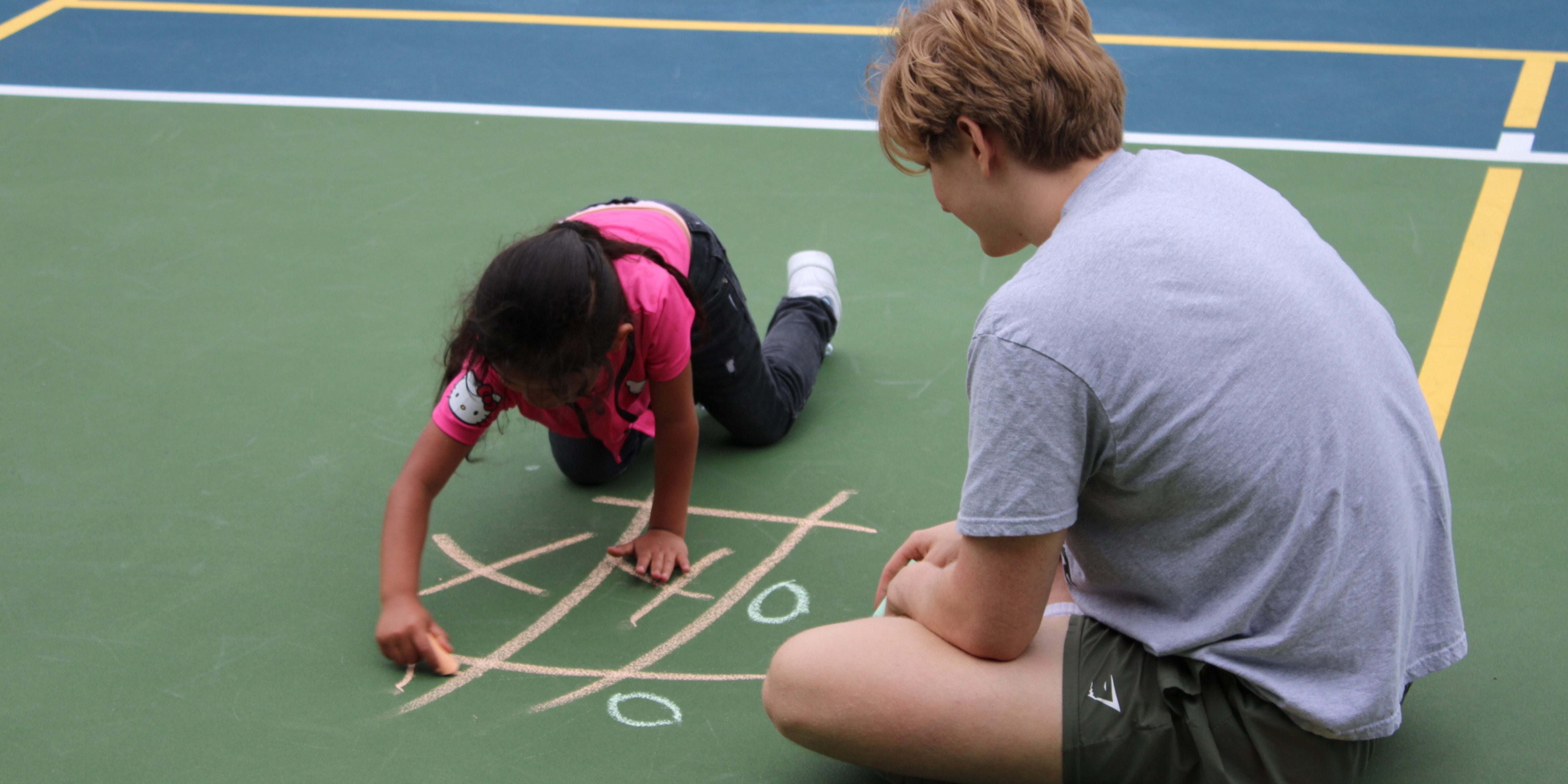 A mentor playing tik tak toe with chalk and a young girl on a tennis court