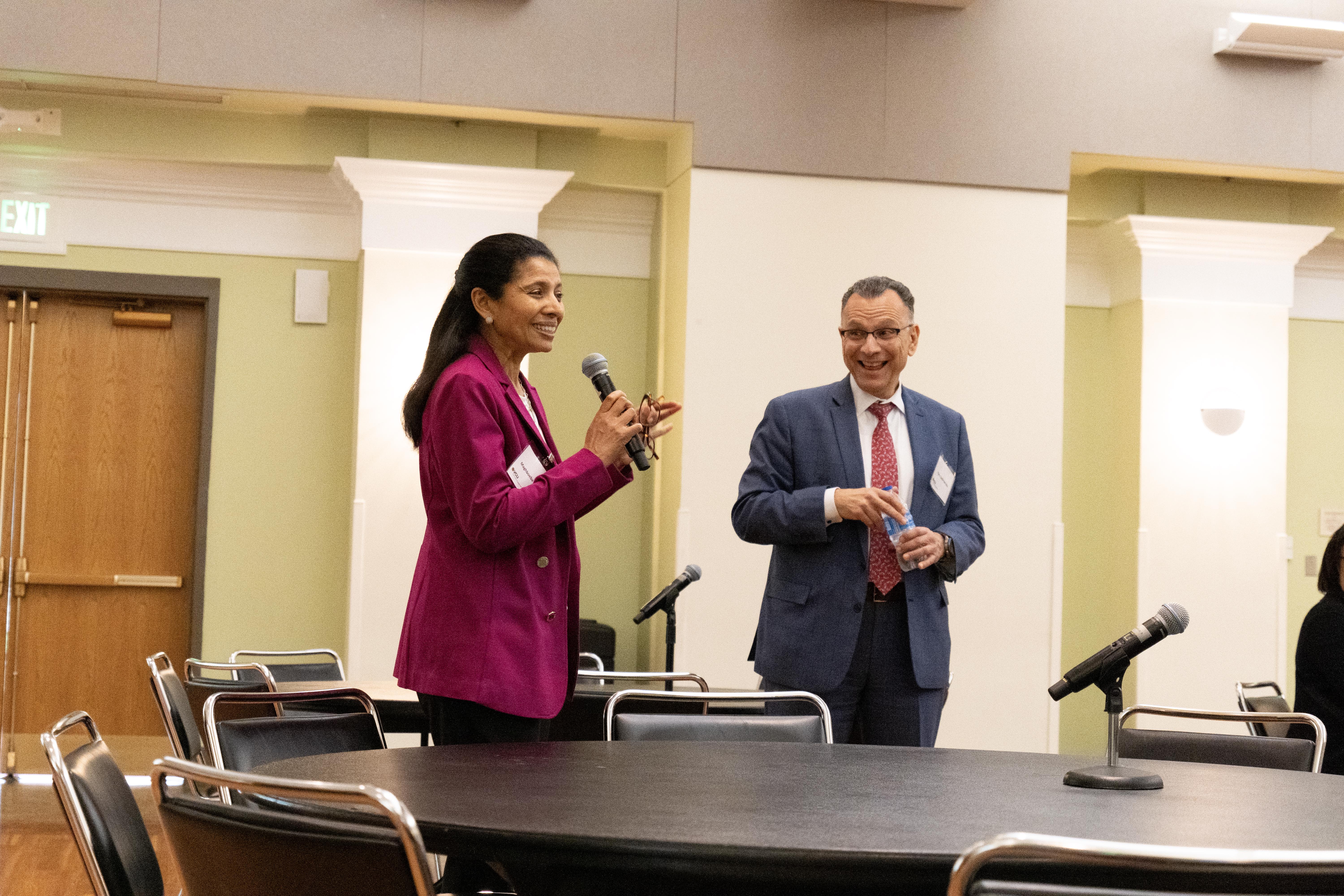 a woman in a pink blazer and a man in a blue suit, engage with the audience at the VCU Council for Community Engagement meeting in May 2024. The woman speaks into a microphone while smiling, and the man gestures with his hand, both standing in front of microphones in a room with chairs and green walls.