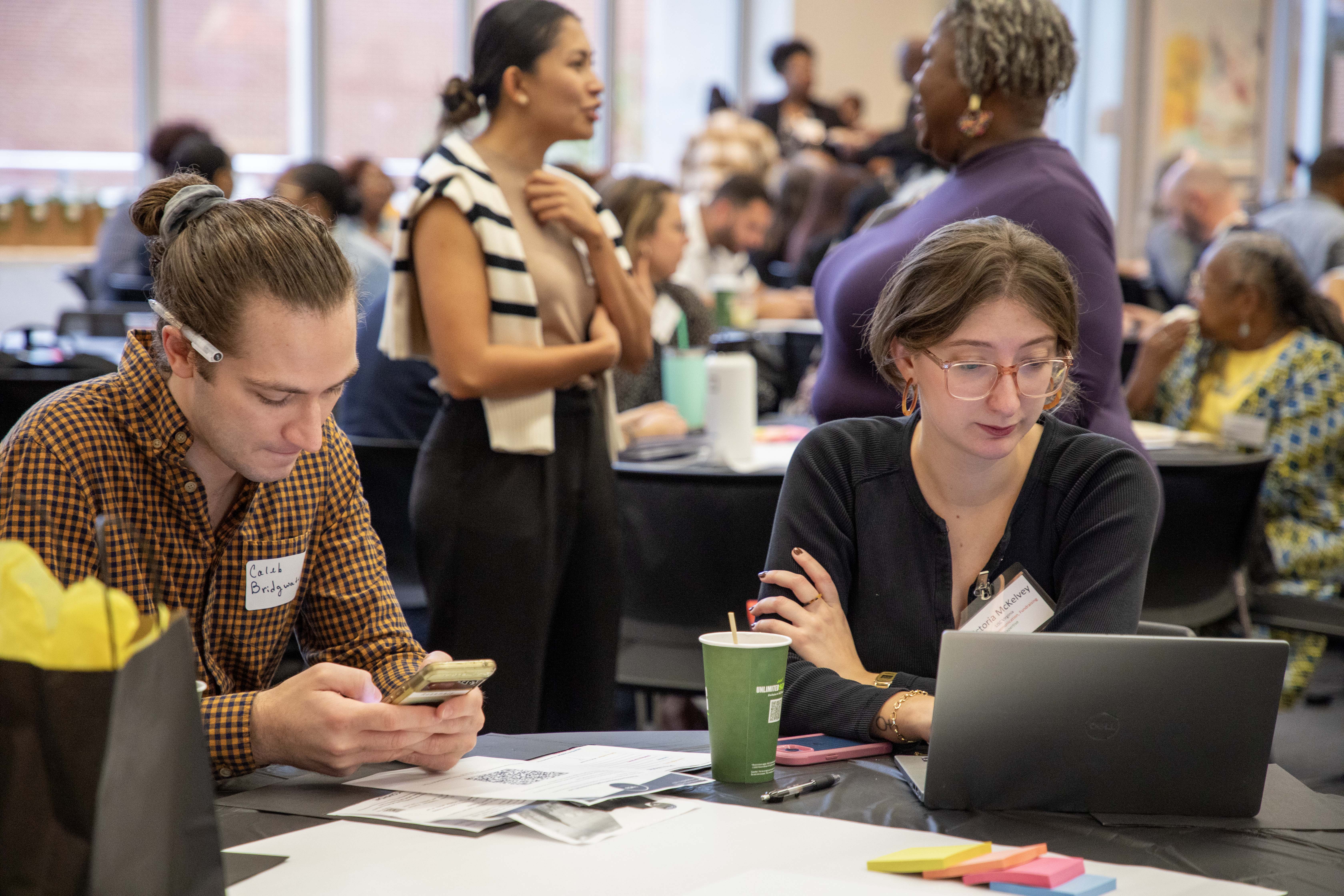 two people people sitting at at table, one person on their laptop the other on their phone with a crowd of people sitting at tables behind them