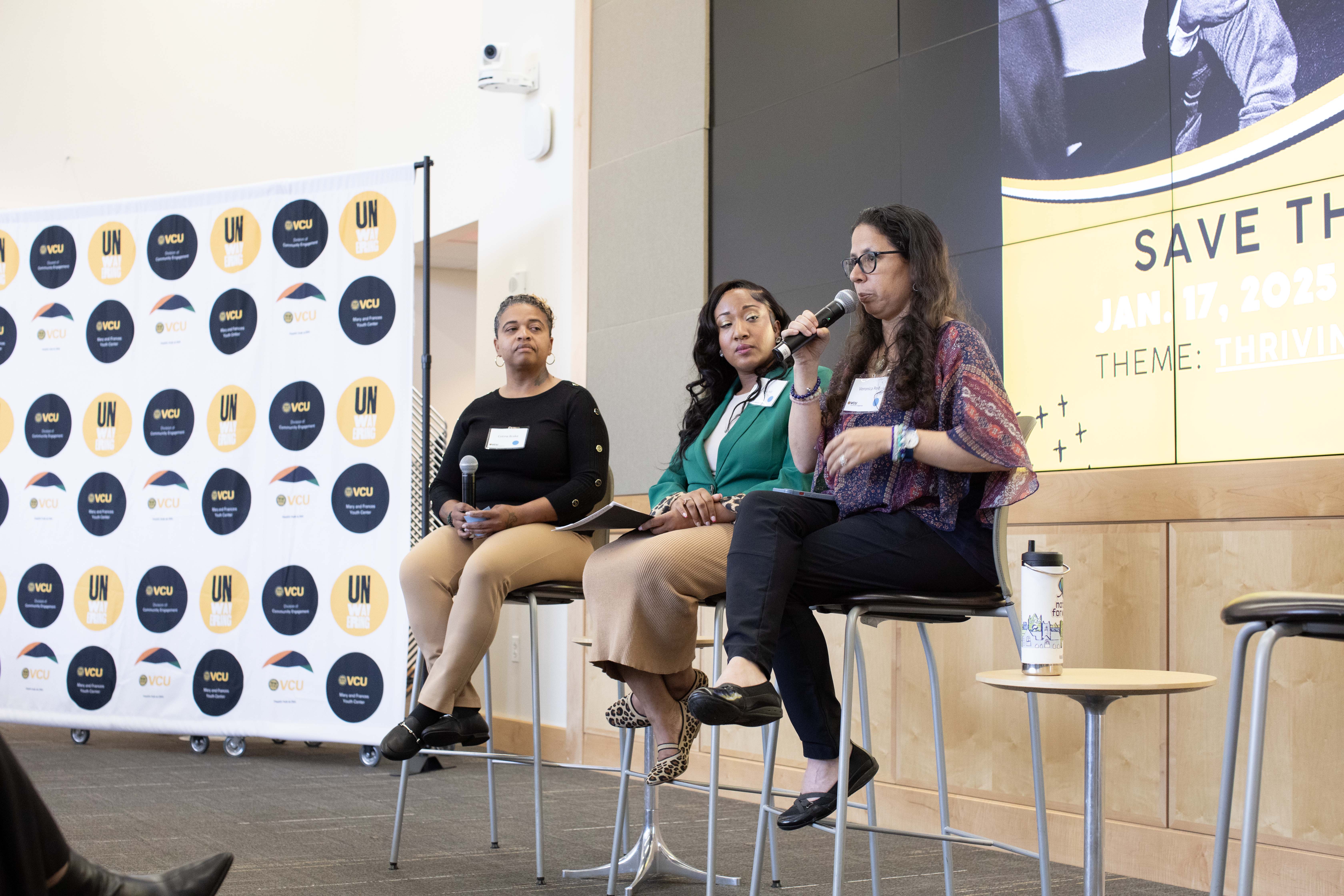 A group of three women panelist sitting in chairs. One woman is speaking in a microphone.