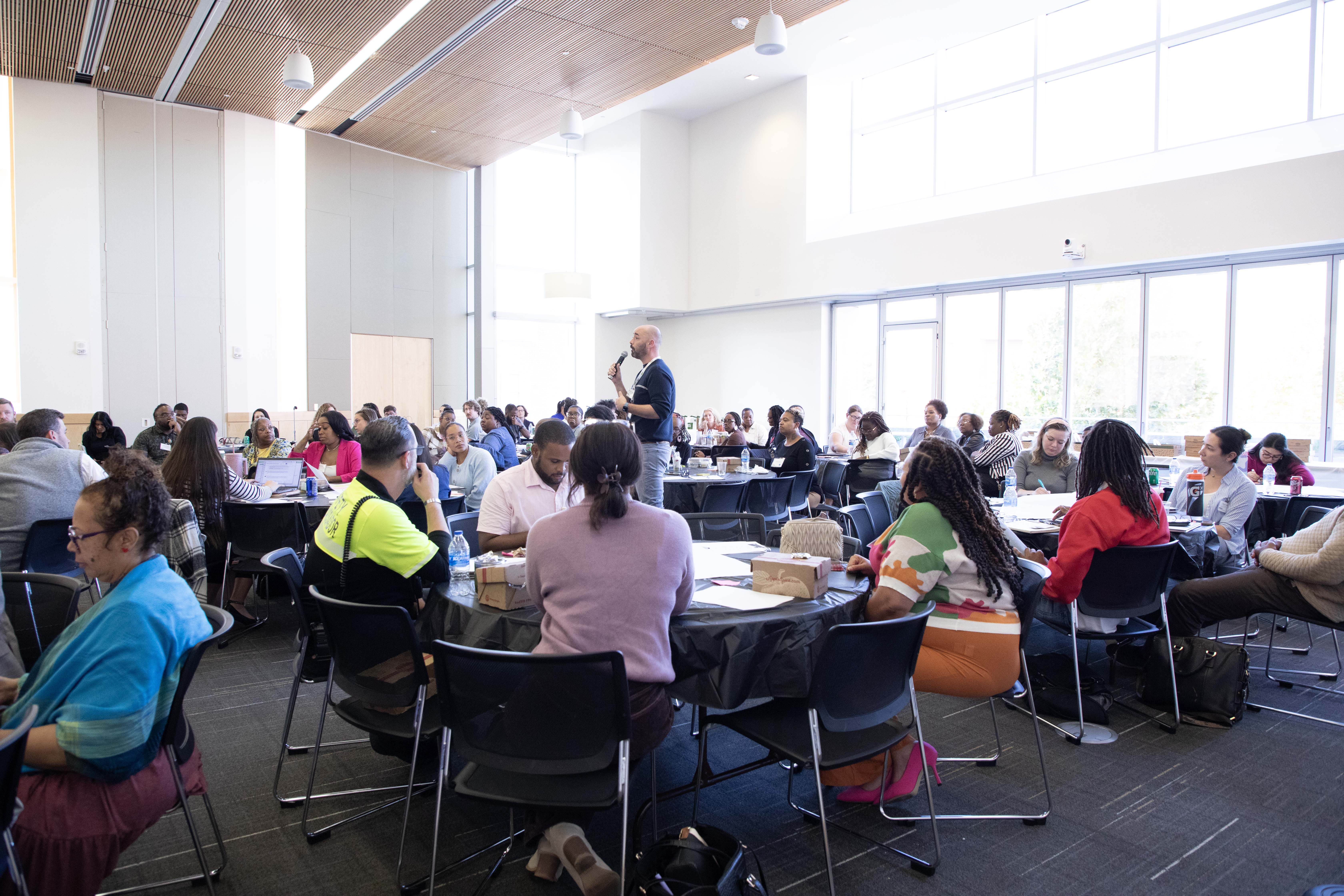 A group of people sitting at their tables in a conference. One man stands speaking into mic